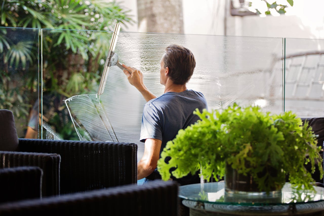 Inicio A man cleaning a glass barrier outdoors with a squeegee, surrounded by greenery.